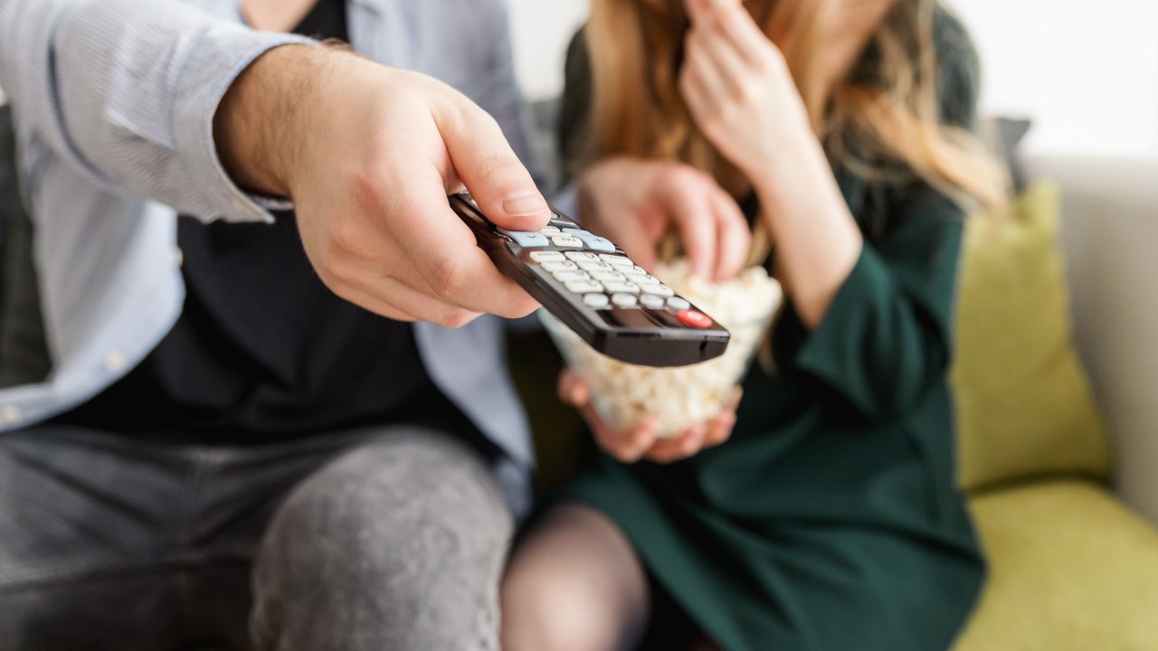 Paar diivanil, üks hoiab kaugjuhtimispulti, teine sööb popkorni